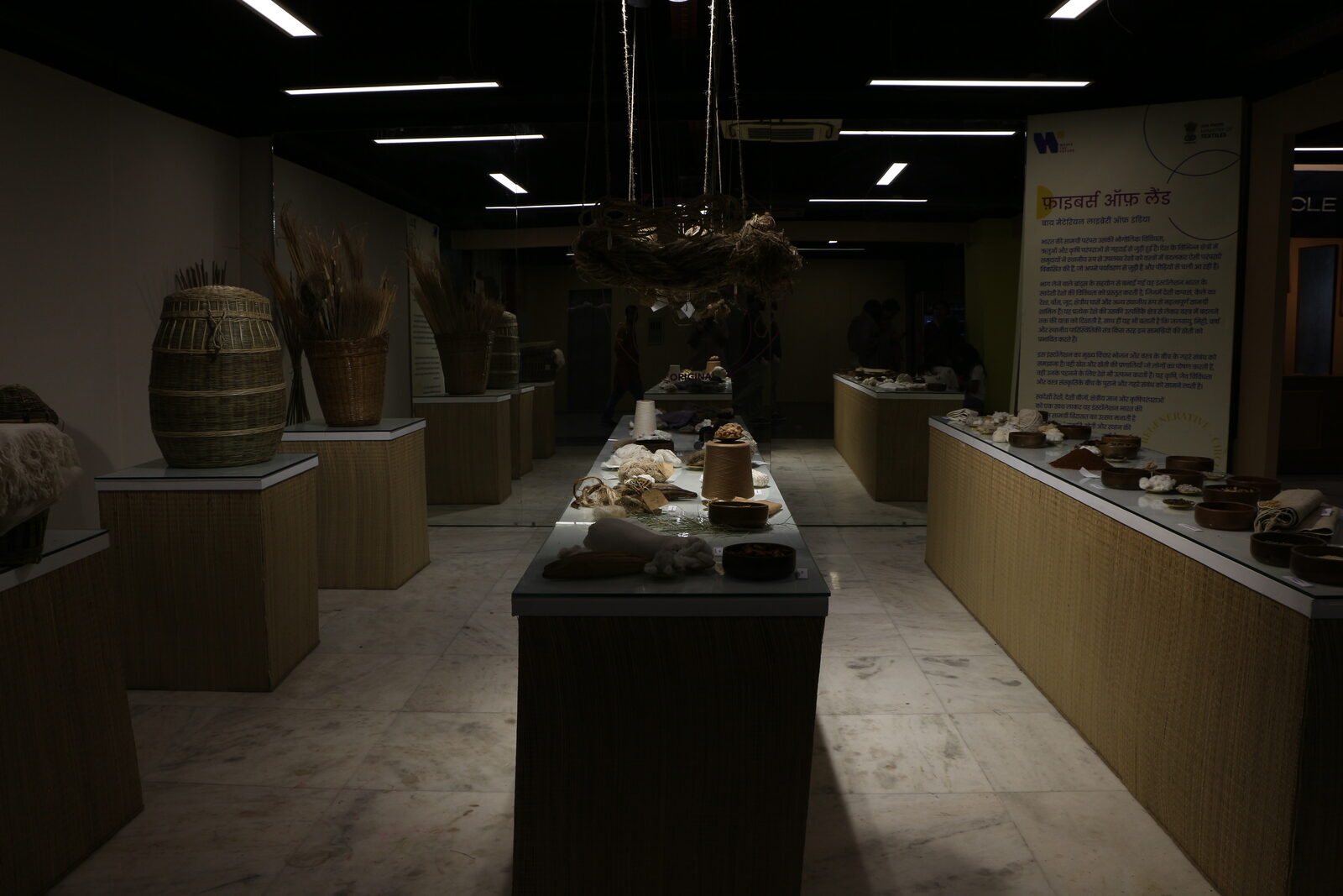 A display gallery at Handloom Haat, Janpath: raw-fibre and craft objects, baskets, spools and bowls, arranged on woven-mat pedestals beneath a hanging rope sculpture, with bilingual wall signage.