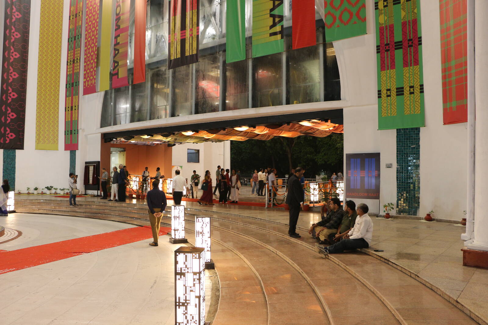The Handloom Haat, Janpath building at night on opening evening: colourful woven-pattern banners hang from the facade above illuminated patterned lantern pillars, visitors and staff arriving along a red carpet.