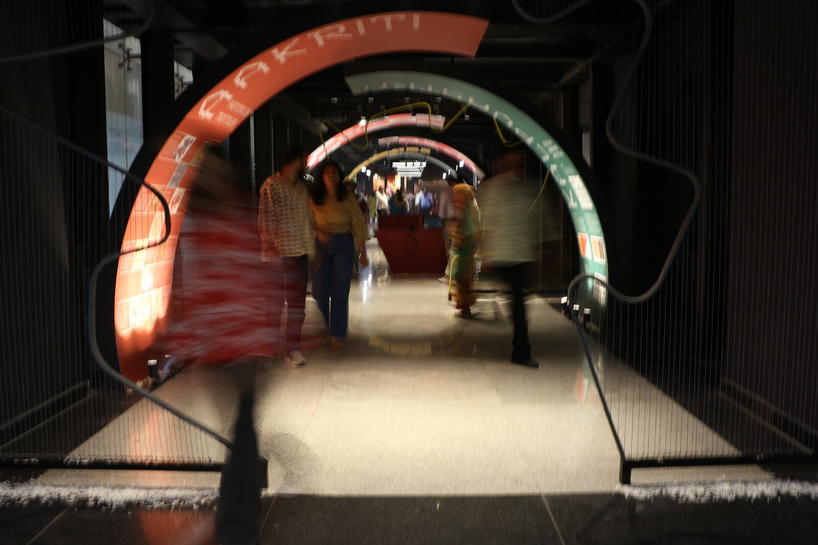 A darkened tunnel installation at Handloom Haat, Janpath: illuminated concentric arches in red and teal with lettering across the beams, visitors walking through in motion toward a lit red desk at the far end.