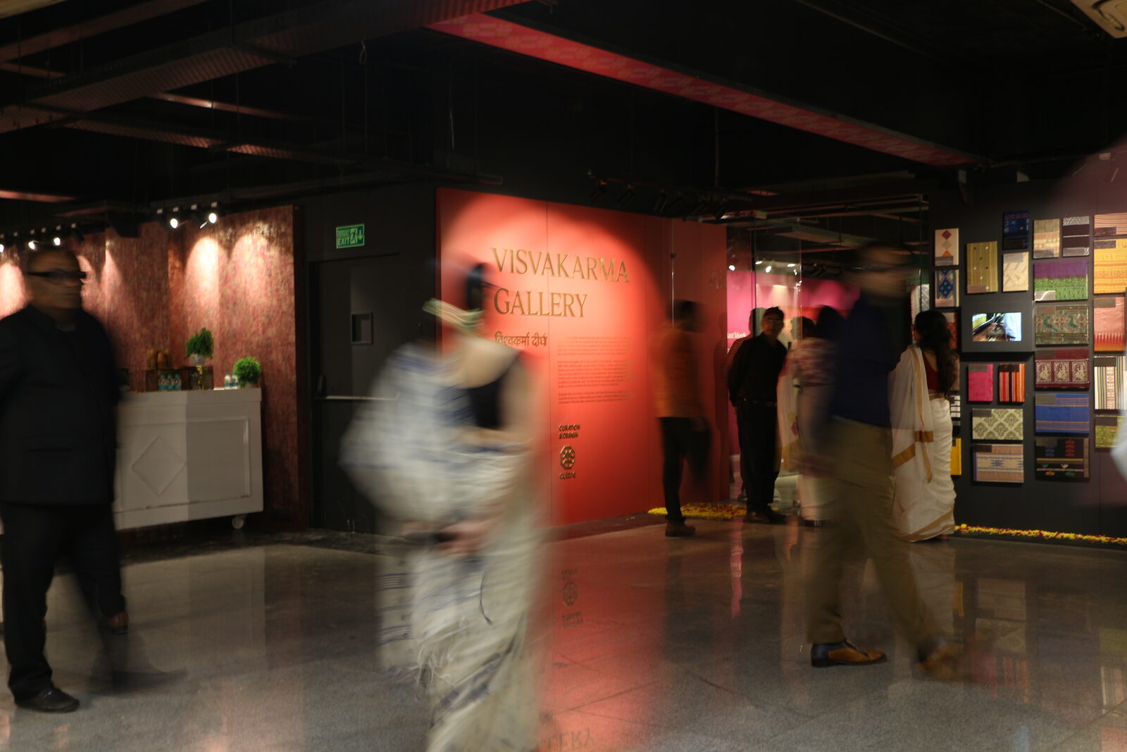 The Vishwakarma Gallery entrance at Handloom Haat, Janpath: a lit red wall reading 'Visvakarma Gallery' beside a wall of framed textile swatches, visitors walking past in motion.